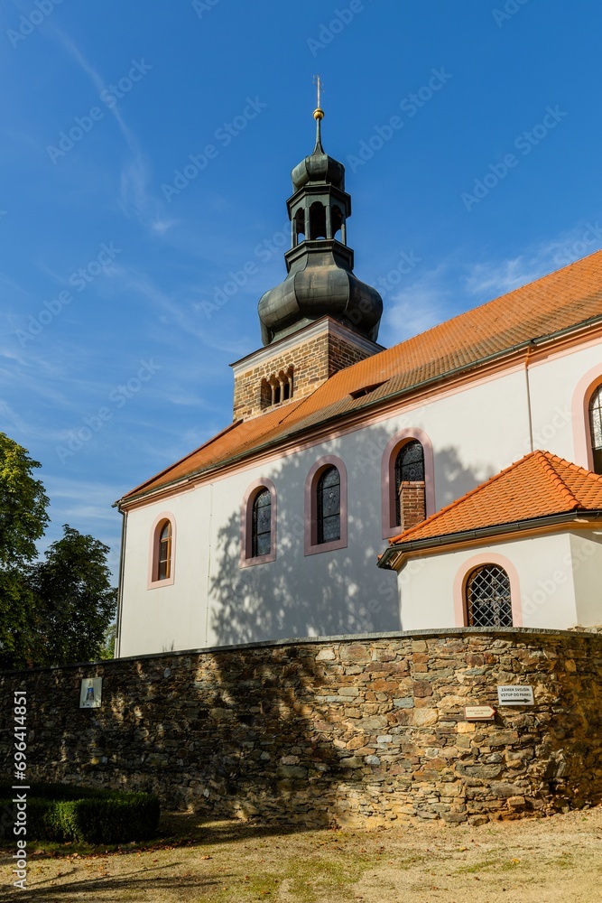Svojsin, Czech Republic - October 13 2023: View of the Saint Petr and Pavel church with a Romanesque stone tower. Dry yellow grass and stone wall in the foreground. Sunny autumn day with blue sky.