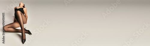 young woman in black bodysuit tights and heel shoes posing on grey background, elegance banner