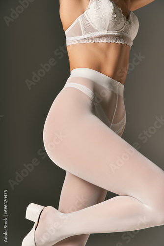 cropped view of model in white lace bra and pantyhose posing on grey background, underwear