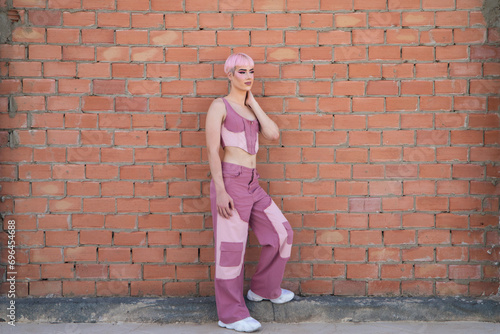 Young gay boy with pink hair and make-up leaning against a brick wall. The boy is dressed modernly in shades of pink. Concept of equality and LGBTQ rights.