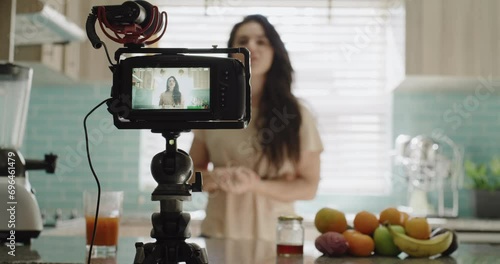 Woman, camera and live streaming in kitchen, blog and tutorial or teaching at home on broadcast. Female influencer, webinar and vlog or technology for video recording, juice and fruit for nutrition photo