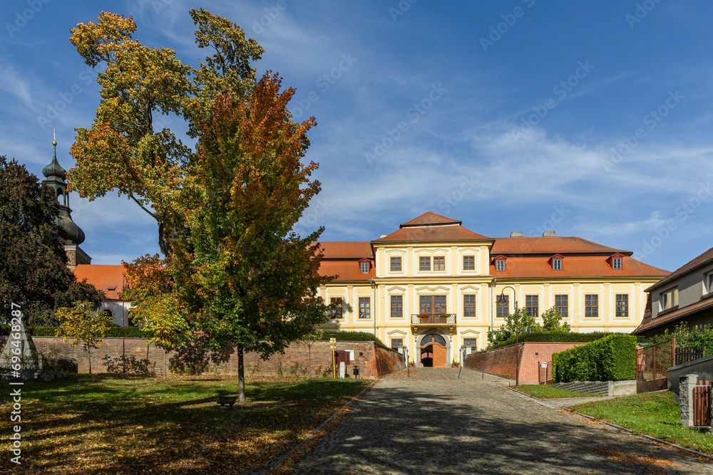 Svojsin, Czech Republic - October 13 2023: Front view of the newly renovated castle, a yellow building with red roof, cobblestone paving, a church tower and a big tree. Sunny autumn day with blue sky.