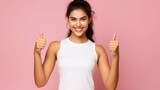 Pretty positive Indian sportswoman wears white cropped top poses with sport equipment holds smartphone points index finger aside on blank space advertises something isolated over pink