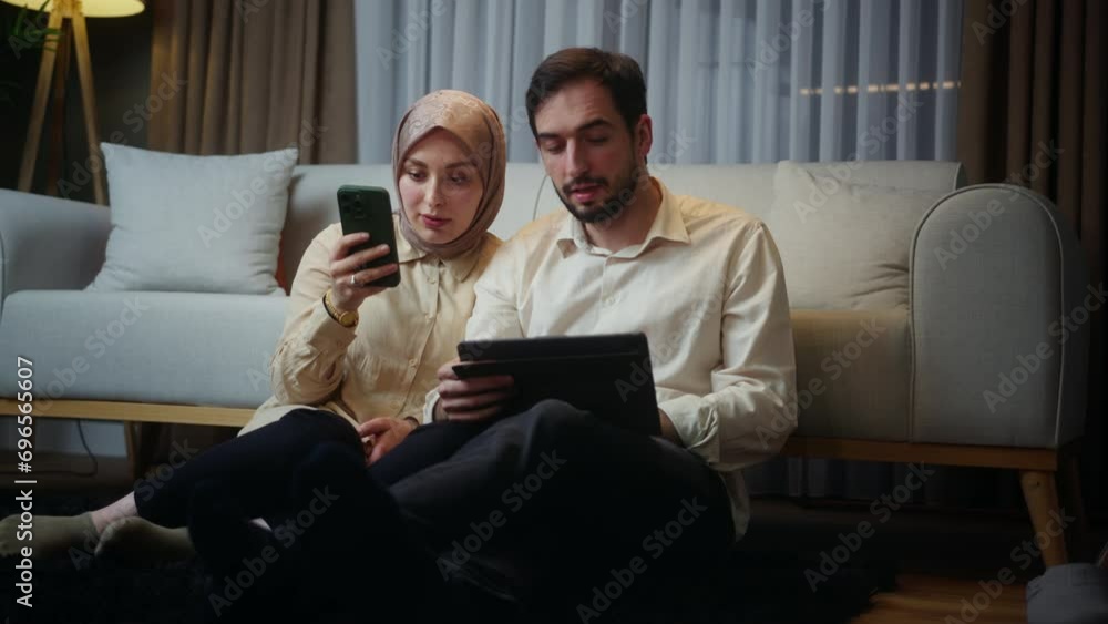 Young married couple leaning against the sofa sitting on floor, woman using smartphone, husband using a tablet device, spending time on internet, using app at home at night Young married couple leaning against the sofa sitting on floor, woman using smartphone, husband using a tablet device, spending time on internet, using app at home at night