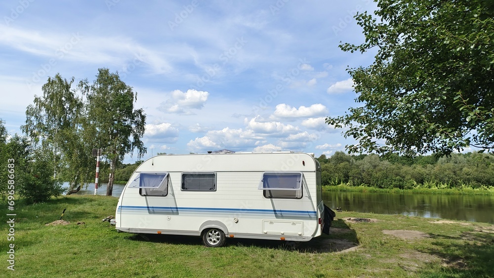 On the grassy bank of the river stands a caravan trailer for a comfortable fishing vacation. There are trees nearby and there is a sign of the ship's passage. On the opposite bank stands a forest