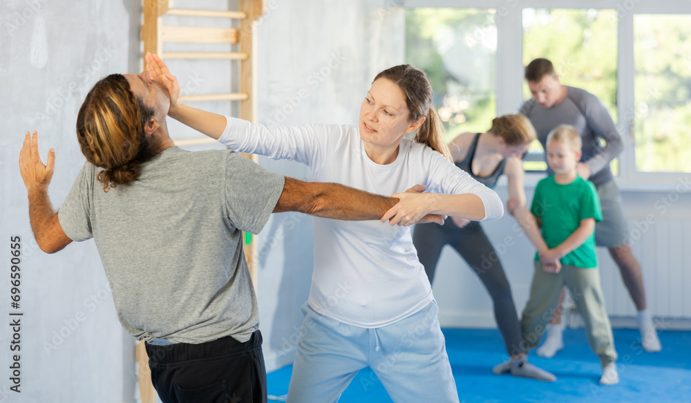 Man and woman at group self-defense lesson are learning new technique of blowing to chin, trainer is practicing technique with children in background