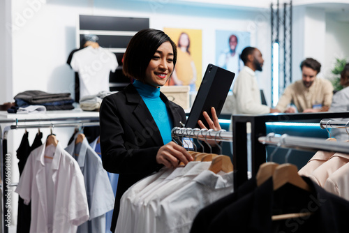 Clothing store smiling manager using digital tablet to check stock availability while browsing through apparel rack. Shopping center boutique asian woman employee managing inventory