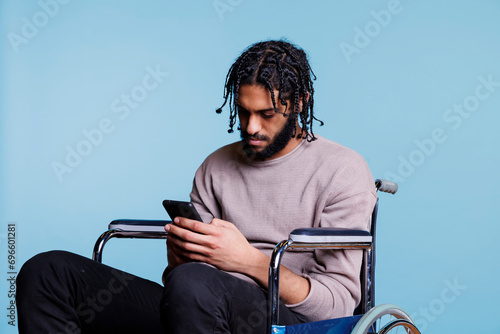 Arab man sitting in wheelchair while scrolling social media on smartphone. Young person with physical impairment sending online message and browsing internet on mobile phone