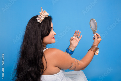 Half-closed portrait of beautiful, black-haired woman against blue background holding and looking into a mirror. photo
