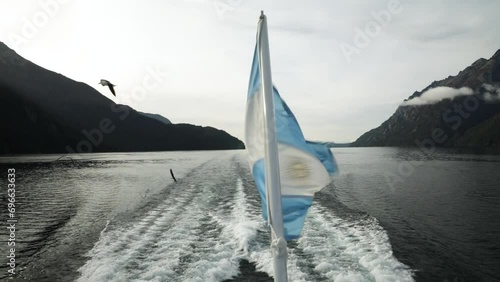 View of the water trail left by the ship sailing Nahuel Huapi at sunset in Bariloche. A flock of seagulls fly around the ship. An Argentinian flag blowing with the wind.  photo