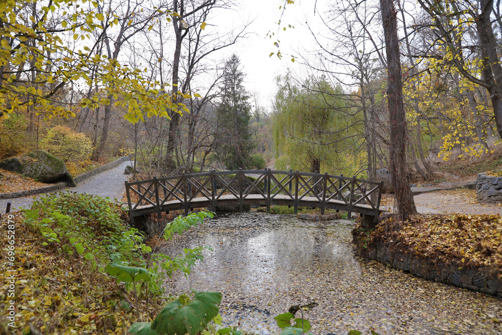 Beautiful Nature Autumn landscape with small bridge. Scenery view on autumn city park with golden yellow foliage in cloudy day. Walking paths in the city Park strewn with autumn fallen leaves Beautiful Nature Autumn landscape with small bridge. Scenery view on autumn city park with golden yellow foliage in cloudy day. Walking paths in the city Park strewn with autumn fallen leaves