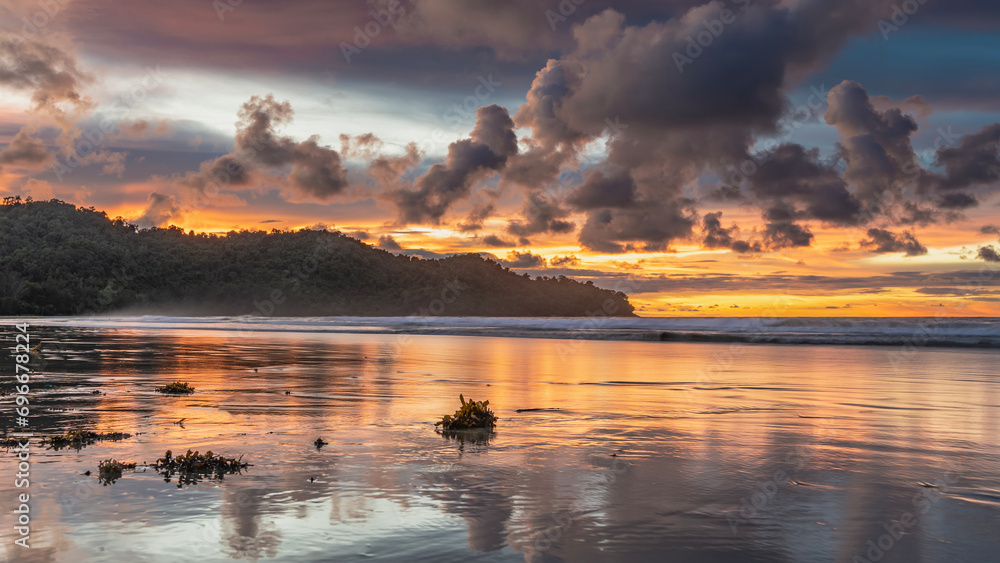 A bright sunset on a tropical island. Purple clouds in the sky illuminated by orange and scarlet. The ocean waves roll towards the shore and spread out on the beach. Reflection on wet shiny sand. 