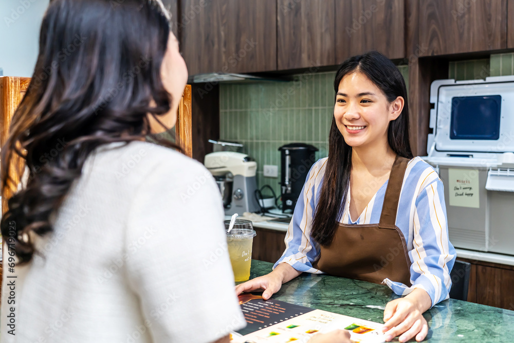 Fototapeta premium Smiling young asian waitress in a cafe with customer for service, payment or order on counter at coffee shop. Serving, employee in small business restaurant helping a girl client at checkout or work