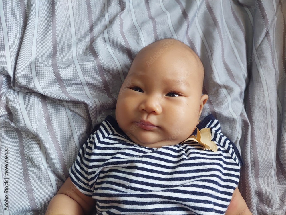 A cute baby with a natural expression wearing a striped shirt, lying on the bed A cute baby with a natural expression wearing a striped shirt, lying on the bed