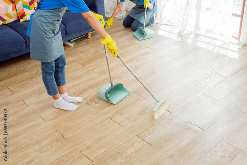 Crop image of a young professional cleaning service women worker working in the house. Girls housekeeper sweeps broomsticks on the wooden floor and cleaning under the sofa. Cleaner and chore concept photo