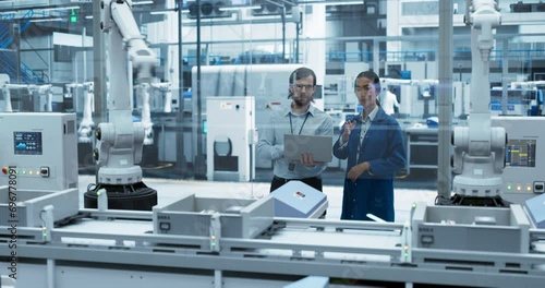 Two Diverse Engineers Standing in a Factory, Using Laptop Computer at an Electronics Factory. Female and Male Specialists Chatting, Monitoring Assembly Robots on a Modern Production Conveyor Belt photo