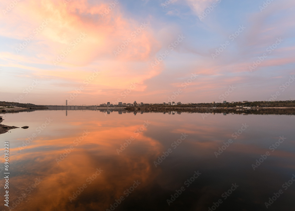 Cityscape during sunset on the bank of a wide river in which the sky is reflected