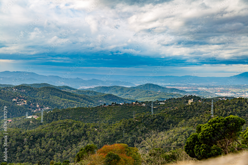 Green leafy wooded mountainous landscape with horizon in background, electric towers, sky covered with abundant thick white clouds with blue tones, cloudy winter day in Barcelona, Catalonia Spain