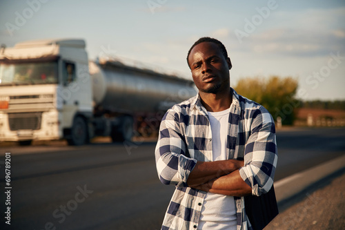 Serious look. With arms crossed. Black man is standing on the road with truck on it © standret