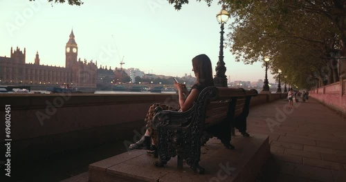 Happy local beautiful female chats via phone, sitting on bench with the view of London famous Big Ben in the evening.