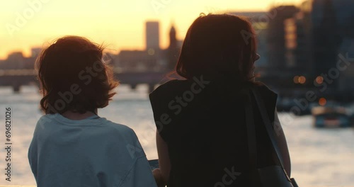 Mother and daughter enjoying beautiful sunset over river together. Aesthetic portrait from behind.