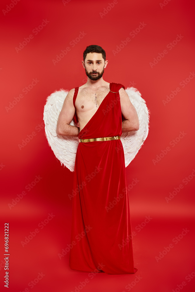 full length of bearded man in cupid costume standing with hands behind back looking at camera on red