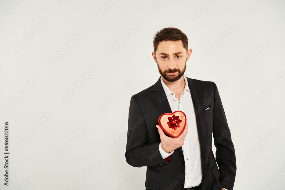smiling charismatic man showing heart-shaped gift box with bow on grey, Saint Valentines concept smiling charismatic man showing heart-shaped gift box with bow on grey, Saint Valentines concept