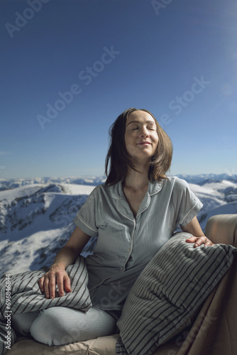 A young beautiful woman in pajamas with her eyes closed in bed against the background of a lake and snowy mountains. A cozy, peaceful atmosphere of nature