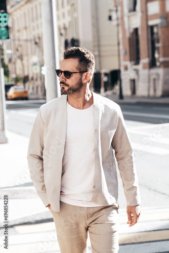 Handsome confident stylish hipster lambersexual model. Sexy modern man dressed in elegant white suit jacket. Fashion male posing in the street background in Europe city at sunset. In sunglasses