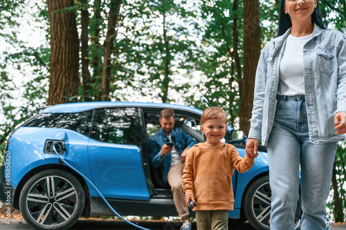 Walking forward. Mother, father and little son are waiting for electric car to charge outdoors photo