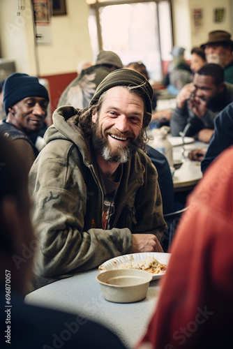 Homeless positive man sits at a table during lunch in a noisy shelter cafeteria, surrounded by other people photo