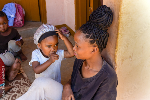 village african mother with braids and her kids in front of the house playing, little girl scratching her head