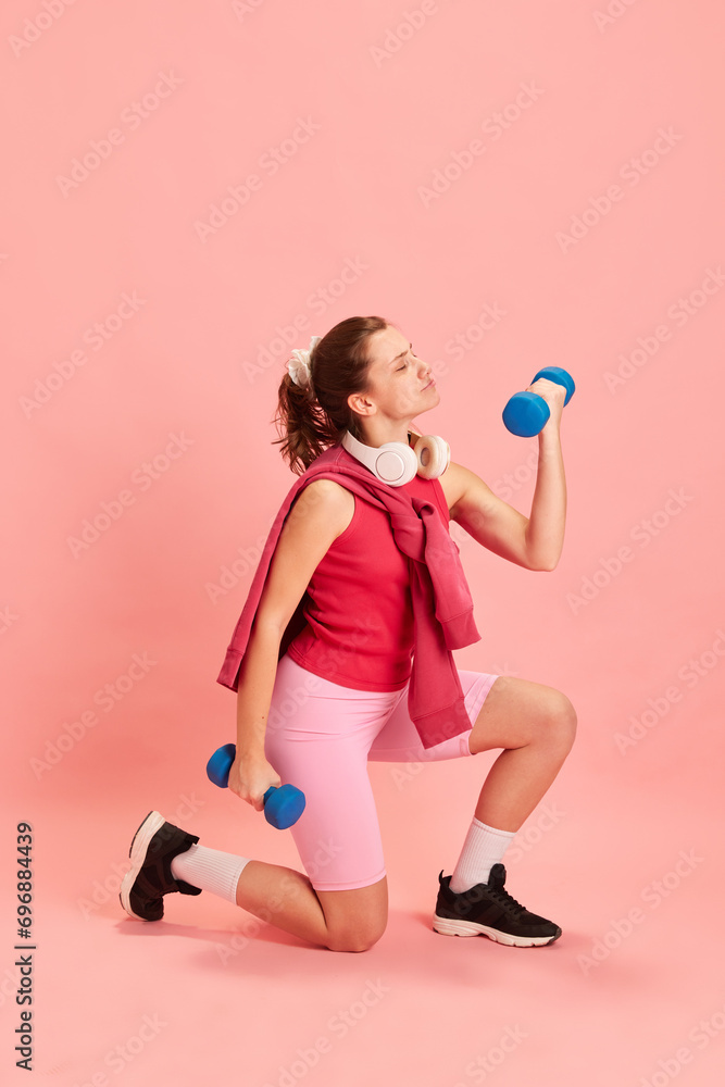 Dynamic portrait of fit young lady in sportwear who exercising with dumbbells against pastel pink background. Concept of sport, active and healthy, lifestyle, athleticism, fitness, energy. Dynamic portrait of fit young lady in sportwear who exercising with dumbbells against pastel pink background. Concept of sport, active and healthy, lifestyle, athleticism, fitness, energy.