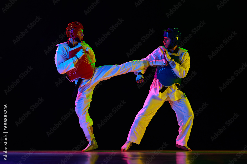 Athletic, strong young men, taekwondo athletes in motion, fighting, training against black background in neon light. Concept of martial arts, combat sport, competition, action, strength