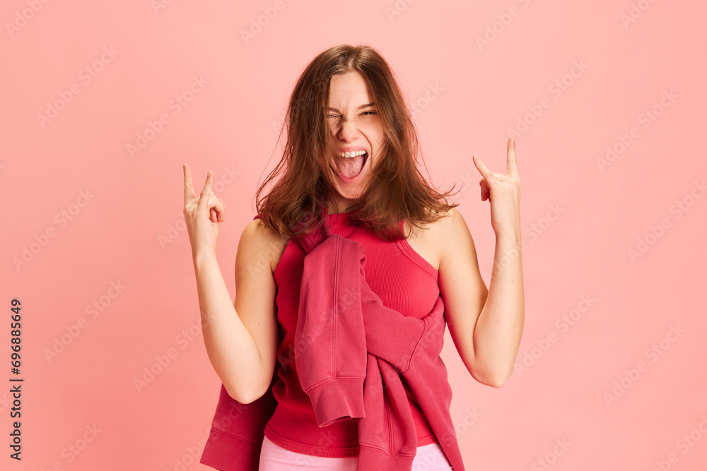 Energetic portrait of young expressive brunette lady dressed sportwear and show rock gesture against pink studio background. Concept of sport, active lifestyle, music, hobby, fitness, energy. Energetic portrait of young expressive brunette lady dressed sportwear and show rock gesture against pink studio background. Concept of sport, active lifestyle, music, hobby, fitness, energy.