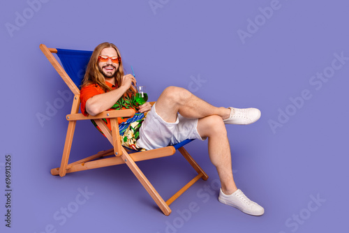 Full length photo of good mood man dressed print shirt lay on deckchair on vacation drink cocktail isolated on purple color background photo