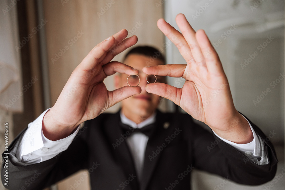 the groom holds the wedding rings with his fingers at the level of his eyes and looks into the camera through the rings. Funny photo. Selective focus.