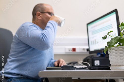 Silhouette of a man drinking from a mug and looking at a computer screen at his workplace.