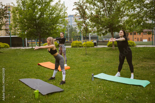 Group of young women does sports exercises on green lawn in park. Trainer shows forward bends to ladies at outdoor workout