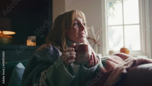 Woman wearing cosy jumper and blanket sitting on sofa at home drinking hot drink from cup - shot in slow motion