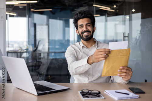 Portrait of a happy young Indian man working in the office, sitting at the desk and opening the received envelope with a letter and documents, smiling at the camera photo