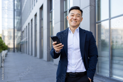 Portrait of a young Asian male businessman standing smiling on the street near an office center, holding a phone and looking at the camera photo