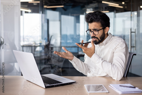 Serious young Indian male businessman sitting in the office at the desk and talking emotionally on the phone through the loudspeaker, using voice search and recording the conversation photo