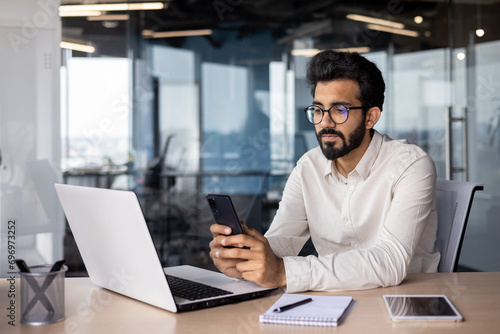 Wall Mural Serious Muslim young man working in office, sitting at desk and using phone, tex