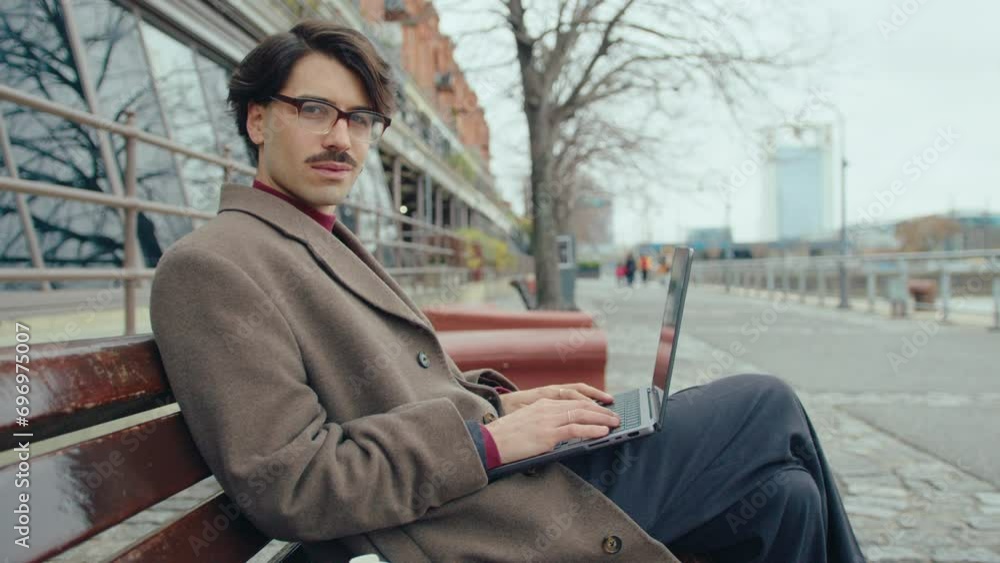 Young handsome businessman in stylish coat and smart casual outfit sitting on bench on city street, holding laptop and posing on camera Young handsome businessman in stylish coat and smart casual outfit sitting on bench on city street, holding laptop and posing on camera
