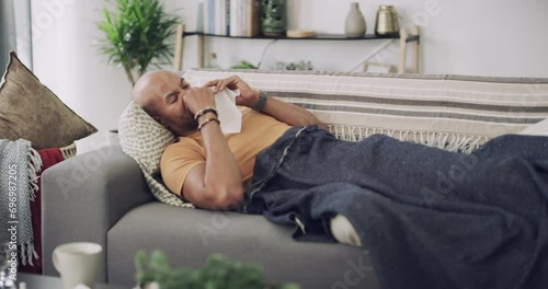 Sick, tissue and blowing nose with black man on sofa in living room of home for recovery, rest or to relax. Cold, flu or allergy and young person sneezing or coughing with blanket for warm comfort photo