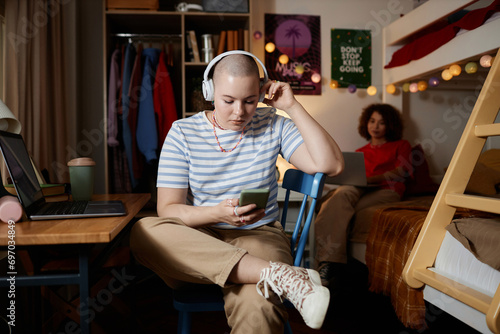 Two female students in college dorm room, focus on gen Z young woman using smartphone in foreground, copy space photo