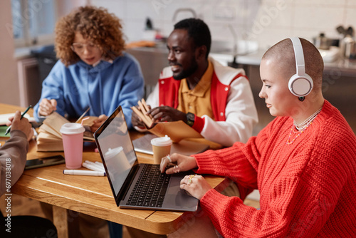 Multiethnic group of young people studying together at table in college dorm.  focus on bald young woman using laptop  copy space