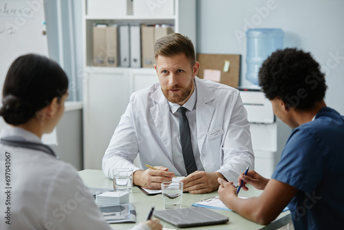Portrait of bearded male doctor wearing lab coat instructing young trainees in meeting photo