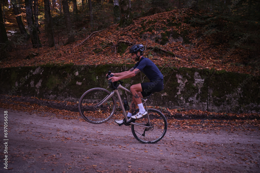 Fototapeta premium Man cyclist on gravel bicycle riding wheelie on a rear wheel in forest. Cyclist practicing on gravel road. Gravel biking during the autumn. Sport motivation. Adventure travel on bike. 
